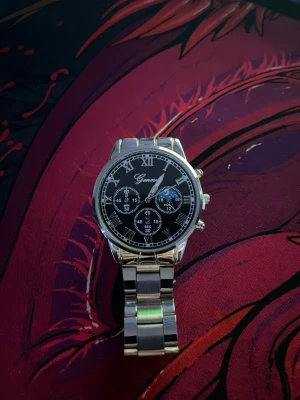 Silverfärgad Geneva klocka med svart urtavla - Stilren klocka från Geneva med silverfärgad länkarmband och svart urtavla. Klockan har romerska siffror, flera dekorativa subdials och en klassisk rund form. Perfekt accessoar för dig som vill ha en tidlös look.