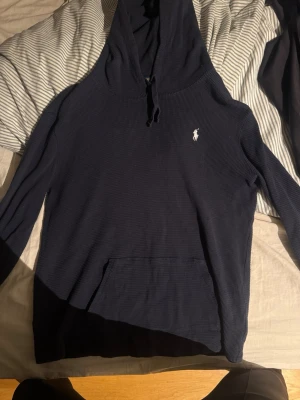 Marinblå hoodie från Polo Ralph Lauren - Snygg marinblå hoodie från Polo Ralph Lauren med klassisk huva och dragsko. Tillverkad i ett ribbat, mjukt material och har en vit broderad logga på bröstet samt en stor magficka framtill. Perfekt för en chill och stilren look.