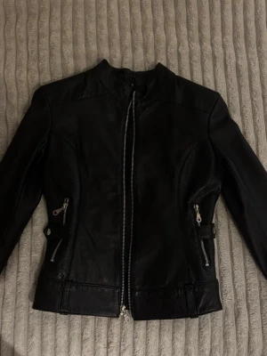 Svart skinnjacka med dragkedjor - Snygg svart skinnjacka i slim fit med perforerade detaljer på sidorna och armbågarna. Jackan har flera silvriga dragkedjor, ståkrage och coola metalldetaljer vid fickorna. Perfekt för dig som gillar en edgy stil och vill ha något som sticker ut.