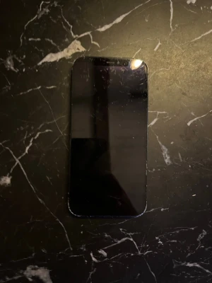 Svart iPhone 12 med dubbla kameror - Säljer en svart iPhone 12 med stilren design och rundade hörn. Telefonen har en slät glasbaksida och två kameror på baksidan tillsammans med blixt. Klassisk Apple-logga på baksidan och metallram runt om. Perfekt för dig som gillar snygg och modern teknik. Väldig andvänd och slite batteri 89 procent det finns 2 fel att bakkameran fungerar bara på 0.5 och touchen där uppe fungerar ibland 