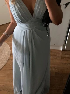 Balklänning - Elegant ljusblå balklänning med djup v-ringning och omlottdesign framtill. Klänningen har tunna axelband och vacker spets med broderier i ryggen. Kjolen är i ett mjukt, flytande tyg som ger en romantisk känsla. Perfekt för bal eller speciella tillfällen. Endast använd 1 gång