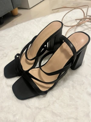 Svarta sandaletter med blockklack - Säljer ett par trendiga svarta sandaletter med fyrkantig tå och höga blockklackar. Sandalerna har tunna remmar i konstläder som korsar foten. Innersulan är beige och lätt vadderad för extra komfort. Aldrig använt de.