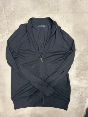 Svart ziptröja från S.C.W. - Säljer en svart ziptröja från S.C.W. med ribbad krage och muddar. Tröjan har hel dragkedja framtill och är tillverkad i ett mjukt, tunt material som passar perfekt till lager-på-lager. Enkel och stilren design som funkar till det mesta.