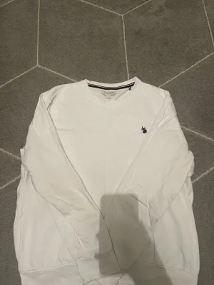 Vit sweatshirt från U.S POLO ASSN. - Klassisk vit sweatshirt från U.S POLO ASSN med rund halsringning och broderad logga på bröstet. Tröjan har långa ärmar och ribbade muddar vid ärmslut och nederkant. Perfekt för en clean och stilren look.
