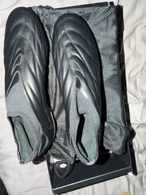 Svarta Adidas Copa Pure+ fotbollsskor - Säljer ett par helsvarta Adidas Copa Pure+ fotbollsskor med strumpliknande skaft och slät ovandel i syntetmaterial. Skorna har en modern, strömlinjeformad design och levereras med skopåse, extra dobbar och verktyg för dobbyte. Perfekta för dig som vill ha stilrena och funktionella skor på planen.