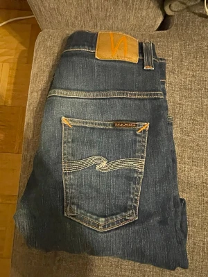 Mörkblå jeans från Nudie Jeans - Säljer ett par mörkblå jeans från Nudie Jeans med klassisk femficksmodell och kontrastsömmar. Jeansen har raka ben och är tillverkade i slitstarkt denim med Nudie-loggan på bakfickan. Perfekta för en avslappnad och stilren look i modell THIN FINN strlk L30, W31