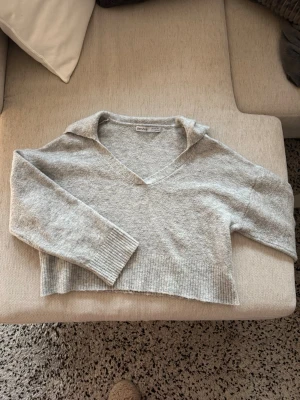 Ljusgrå croppad stickad tröja Bershka - Ljusgrå croppad stickad tröja från Bershka med v-ringning och krage. Tröjan har långa ärmar och bred ribbstickad kant nedtill och vid ärmsluten. Perfekt för lager på lager och en chill look.