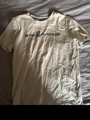 Beige t-shirt från Sail Racing - Snygg beige t-shirt från Sail Racing med svart logotyptryck på bröstet. Klassisk rund halsringning och korta ärmar. Tillverkad i mjuk bomull som känns skön mot huden. Perfekt för en avslappnad och stilren look.