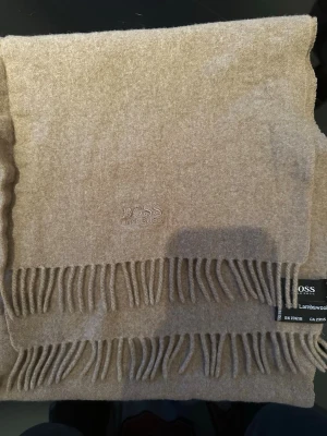 Beige halsduk från Hugo Boss - Snygg beige halsduk äkta Hugo Boss i mjuk lammull. Halsduken har klassiska fransar i kanterna och diskret broderad logga framtill. Perfekt accessoar för att höja din outfit med en stilren touch.