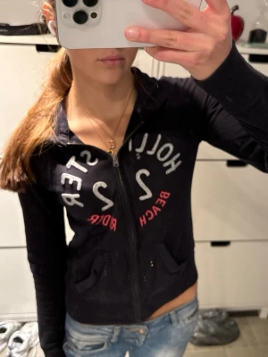 Hollister zip up hoodie huvtröja med dragkedja - Snygg mörkblå nästan svart zip hoodie från Hollister med vit och rosa text på bröstet.                     Storlek XS!                                                            Hoodien har dragkedja framtill, två fickor och långa ärmar. Perfekt till jeans för en avslappnad look.           Dragskon fattas men annars bra skick!