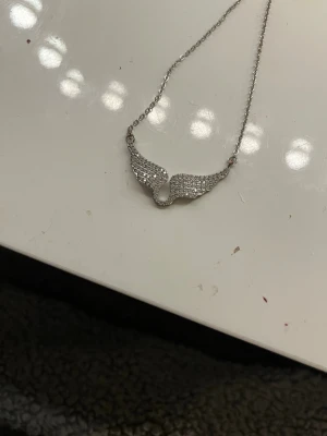 Silverhalsband med gnistrande vingar - Snyggt halsband i silverfärg med ett hänge format som två vingar täckta av små glittrande stenar. Kedjan är tunn och elegant, och vingarna ger ett coolt och unikt intryck. Perfekt för dig som gillar detaljer och vill sticka ut lite extra.