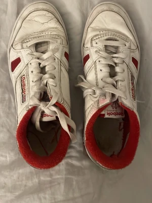 Vita Reebok Classic sneakers med röd kant nypris 1100kr - Säljer ett par vita Reebok Classic sneakers med röda detaljer och röd vadderad kant. Skorna har Reebok-logga på sidan och bak, samt klassisk snörning. Tillverkade i läder med retrovibe och låg profil. Perfekta för dig som gillar old school-stil.