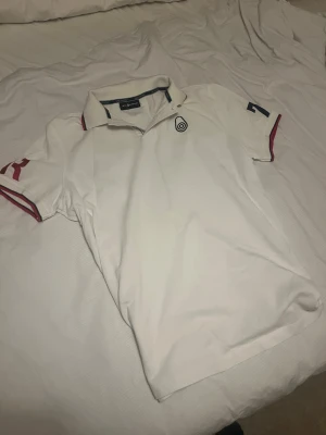 Vit pikétröja från Sail Racing - Klassisk vit pikétröja från Sail Racing med krage och knäppning framtill. Tröjan har röda och blå detaljer på ärmarna samt en broderad logga på bröstet. Perfekt till en sportig och avslappnad stil.