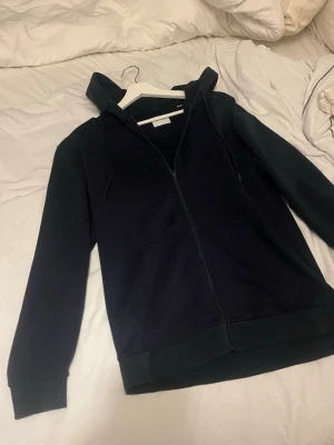 Svart hoodie med dragkedja Jack & Jones - Snygg svart hoodie från Jack & Jones med dragkedja framtill och två fickor. Klassisk modell med huva och justerbara snören. Perfekt för en avslappnad stil och enkel att matcha med jeans eller joggers.