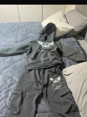 Grått loungewear-set med tryck - Mysigt grått loungewear-set med hoodie och byxor. Setet har tryck med fågelmotiv och text i vitt och blått på både tröja och byxa. Byxorna har praktiska fickor med kardborre. Perfekt för chill dagar hemma.