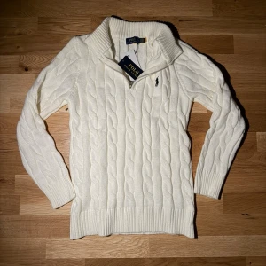 Vit Zip-Knit Polo Ralph Lauren - Kabelstickad vit tröja från Polo Ralph Lauren med klassisk polokrage och svart broderad logga på bröstet. Tröjan har långa ärmar, ribbade muddar och är gjord i mjukt material som känns lyxigt och bekvämt.TVEKA INTE ATT SKICKA EN BUD:)
