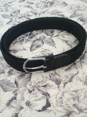 Svart flätat bälte med silverfärgat spänne - Snyggt svart flätat bälte med silverfärgat metallspänne och detaljer i skinn. Perfekt accessoar för att lyfta din outfit och passar till både jeans och byxor. Enkel och stilren design som funkar till det mesta.