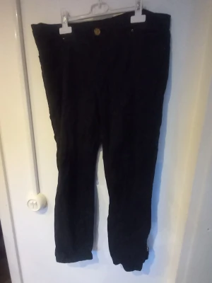 Svarta byxor med dragkedja vid benslut - Säljer ett par svarta byxor med raka ben och dragkedjor vid bensluten. Byxorna har knappstängning och ser ut att vara gjorda i ett mjukt tyg, troligen bomull eller bomullsblandning. Perfekta för dig som gillar en enkel men ändå cool stil.