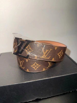 Brunt LV bälte - Snyggt brunt skärp från LV med klassiskt monogrammönster och svart LV-spänne. Tillverkat i skinn med beige insida och detaljer i guldbrunt. Perfekt accessoar för att lyfta din outfit.110CM verktyg för att justera storlek följer med 