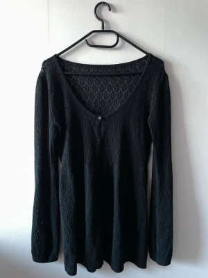 Svart spetsmönstrad kofta - Snygg svart kofta med spetsmönster och djup urringning. Koftan har långa ärmar och knäpps med små svarta knappar framtill. Materialet är tunt och genombrutet.