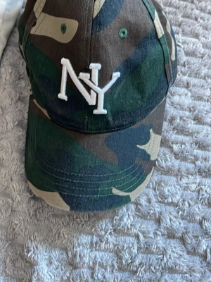 Camo keps från Statewear NY - Säljer en camouflagemönstrad keps från Statewear med broderad vit NY-logga framtill. Klassisk baseballmodell med böjd skärm och justerbar velcro baktill. Tillverkad i bomullsmix och perfekt för dig som gillar streetwear och sportig stil.