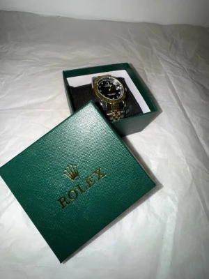 guld och silver klocka - Snygg klocka med svart urtavla, guldfärgad räfflad ring och tvåfärgad länk i guld och silver. Klockan har datumvisning och markerade index med stenar. Kommer i original grön box.