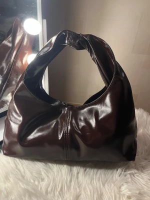 Mörkbrun handväska - Snygg mörkbrun handväska i glansigt fejkläder med mjuk, slouchy form och bred axelrem. Väskan har minimalistisk design utan synliga loggor och passar perfekt till en trendig look. Perfekt storlek för det viktigaste och ger en cool vibe till outfiten.