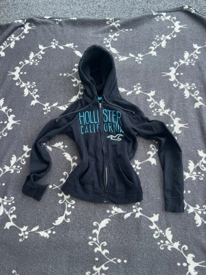 Marinblå Hollister hoodie Dam - Snygg marinblå hoodie från Hollister med turkosa detaljer och texten 'Hollister California' broderat på bröstet. Strl XS. Finns en liten defekt som man ser på bilden men den är väldigt liten. Kolla in min profil för mer kläder