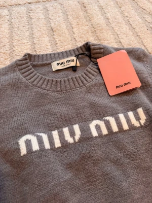Grå stickad tröja från Miu Miu - Supersnygg grå stickad tröja från Miu Miu med vit logotyp framtill. Tröjan har rund halsringning, långa ärmar och ribbade muddar vid ärmslut och nederkant. Perfekt för dig som gillar stilrena och trendiga plagg.