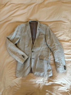 Ljusgrå linnékavaj från tiger of sweden - Snygg ljusgrå kavaj i linne med två knappar framtill och klassiska fickor. Kavajen har smal passform, slits bak och dekorativa knappar vid ärmsluten. Perfekt för en stilren och avslappnad look för festen! Nyprs 4999:-