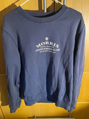 Mörkblå sweatshirt från Morris - Snygg mörkblå sweatshirt från Morris med tryck på bröstet 'Gentlemens Club'. Tröjan har rund hals, ribbade muddar och är långärmad. Materialet är mjuk bomull som känns skönt mot huden. Perfekt för dig som gillar klassisk och stilren design.
