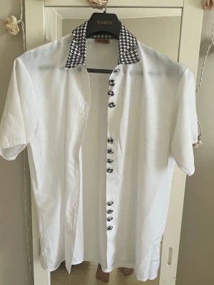 Vit skjorta  - Vit skjorta    med korta ärmar och en svartvit hundtandsmönstrad krage. Skjortan har knappar framtill och är tillverkad i ett lätt bomullstyg. Perfekt för dig som vill ha en unik twist på en klassisk look.