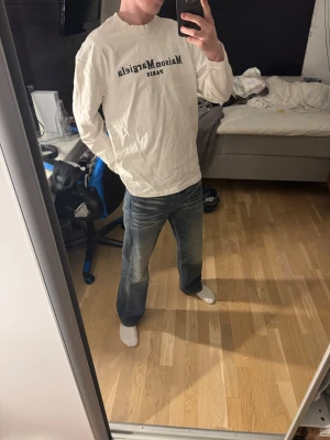 Maison Margiela vit långärmad tröja - Säljer en vit långärmad tröja från Maison Margiela med svart tryck framtill där det står 'Maison Margiela PARIS'. Tröjan har avslappnad passform och rund halsringning. Perfekt för dig som gillar stilrena och exklusiva plagg.