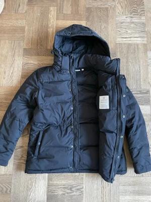 Svart dunjacka med luva, dam: Norrsken - Klassisk svart dunjacka från Norrsken med luva och dragkedja framtill. Jackan har två sidofickor med knappar och innerficka. Den är helt oanvänd i nyskick. Varm vinterjacka dam eller unisex. Stl 36. 