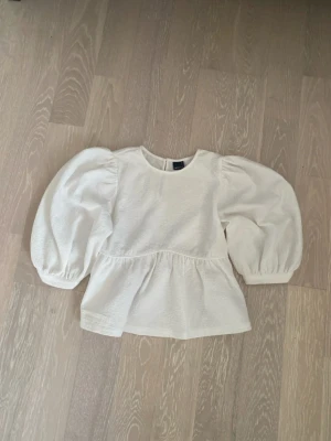 Vit blus med puffärmar - Trendig vit blus med rund hals och stora puffiga långärmar. Blusen har en lätt strukturerad bomullskänsla och en volang nedtill som ger en söt siluett. Perfekt för dig som gillar en stilren men ändå lekfull look.