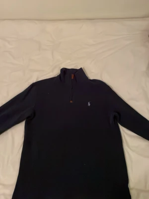 Mörkblå halvzip från Ralph Lauren - Klassisk mörkblå tröja från Ralph Lauren med halv dragkedja och broderad logga på bröstet. Tröjan har hög krage och långa ärmar, perfekt för lager på lager. Snygg och tidlös design som funkar till det mesta.