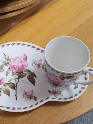 Blommigt kopp- och fatset i porslin - Säljer ett fint kopp- och fatset i vitt porslin med romantiskt ros-motiv i rosa och grönt. Helt nytt!! Både koppen och fatet har matchande blommönster och dekorativa blad runt kanterna. Perfekt för te eller kaffe och ger en klassisk känsla till fikastunden.