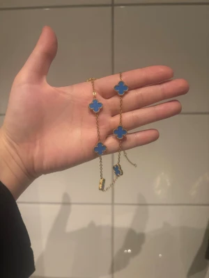 Guldhalsband med blå klöverhängen - Snyggt halsband i guldton med flera blå fyrklöverformade hängen. Kedjan är tunn och elegant, och varje hänge har en pärlad kant som ger extra detalj. Perfekt för dig som gillar färg och unika smycken.