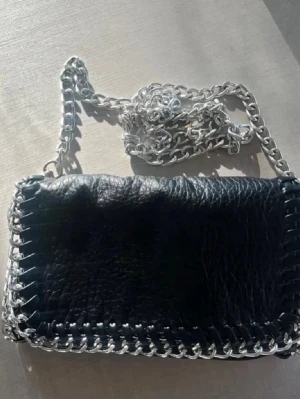 Svart axelväska med kedja - Säljer en svart axelväska i skinnimitation med grov silverfärgad kedja, minsta storleken från tiamo❤️