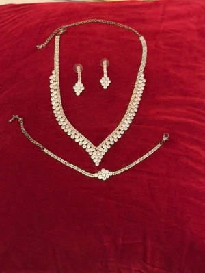 Glittrande smyckesset med strass - Säljer ett elegant smyckesset med halsband, armband och örhängen i silverfärg med gnistrande strassdetaljer. Perfekt för att lyfta din outfit med extra bling. Halsbandet har V-formad design och örhängena är droppformade. Materialet är metall och strass.