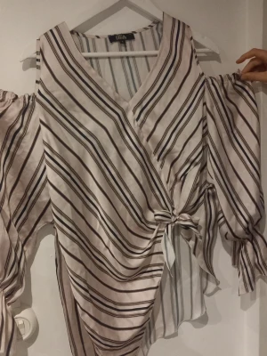 Randig omlottopp från Briefly - Säljer en snygg randig omlottopp från Briefly i ljusa toner. Toppen har v-ringning, knyt i sidan och långa ärmar med resår vid handlederna. Perfekt för dig som gillar en avslappnad men trendig stil.