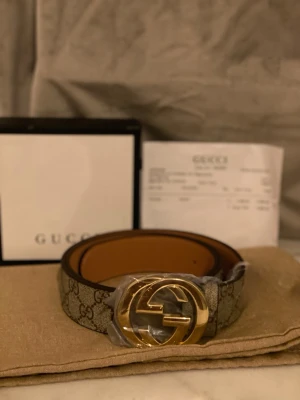 Gucci GG bälte - Snyggt Gucci GG Supreme bälte med klassiskt monogrammönster och stor guldfärgad GG-logga som spänne. Bältet är beige och brunt med exklusiv känsla och passar perfekt till jeans eller byxor för en trendig look.