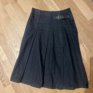 Mörkblå denim  midikjol med bälte stl XS - Snygg mörkblå midikjol  i denim-look med markerade veck och ett dekorativt bälte i midjan. Kjolen har en klassisk A-linjeform och stängs med dragkedja i sidan. Perfekt för dig som gillar stilrena och tidlösa plagg.