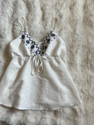 Vit broderad linnetopp med knyt - Söt vit linnetopp med tunna axelband och blå broderade blommor vid urringningen. Toppen har knyt i midjan och är tillverkad i ett lätt, strukturerat tyg som passar perfekt till sommaren. Perfekt för en avslappnad och trendig look.