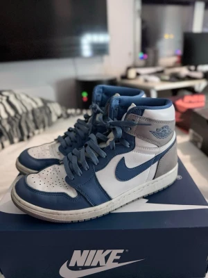 Nike Air Jordan 1 High blå/vit/grå - Säljer ett par Nike Air Jordan 1 High sneakers i blått, vitt och grått. Skorna har klassisk hög siluett, blå swoosh och detaljer, samt grå mocka på hälen. Ovandelen är i läder och mocka, med perforerad tåbox och Air Jordan-logga på sidan.