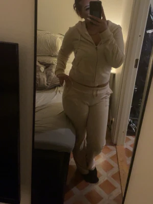 Juicy set,Beige mjukisdress med dragkedja - Dock urtvättad, Säljer en beige mjukisdress med hoodie och dragkedja framtill samt matchande byxor med resår i midjan och snörning. Perfekt för chill dagar hemma. Mjuk och skön kvalitet, troligtvis i bomull eller polyesterblandning.
