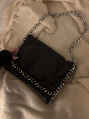 Svart axelväska med kedjedetaljer - Snygg svart axelväska från TIAMO med silverfärgade kedjor som axelrem och runt kanterna. Väskan har magnetlås och ett innerfack med dragkedja. 