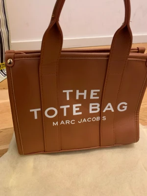 Brun The Tote Bag från Marc Jacobs - Snygg brun handväska från Marc Jacobs med texten 'The Tote Bag' tryckt i vitt på framsidan. Väskan är tillverkad i slätt läder och har både handtag och avtagbar axelrem. Invändigt finns en ficka och dragkedja. Väskan är helt oanvänd, är inköpt utomlands. Vid frågor skriv gärna!