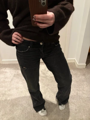 Svarta jeans med slitning - Säljer ett par svarta jeans med låg midja och avslappnad passform. Jeansen har en liten slitning vid ena benet och klassiska fem fickor. Perfekta för en chill och trendig look.