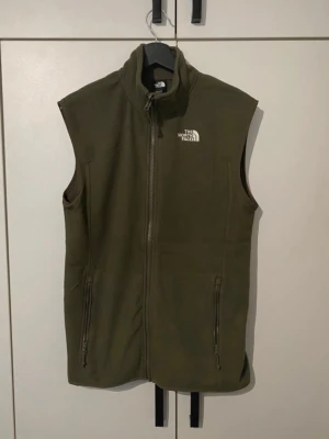 Olivgrön fleeceväst från The North Face - Säljer en olivgrön fleeceväst från The North Face med dragkedja framtill och två fickor med dragkedja. Västen har hög krage och logga broderad i vitt på bröstet. Perfekt för lager-på-lager och outdoor-stil.